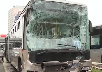 Vista frontal de un bus gris del Metropolitano con el parabrisas delantero completamente roto y la parte inferior del parachoques dañada