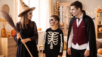 Una mujer con sombrero y escoba de bruja, un niño con maquillaje de calavera y un hombre con capa de vampiro permanecen de pie en una sala.