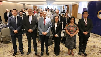 Personas sentadas en mesas redondas con manteles azules dentro de un salón con alfombra y sillas grisáceas