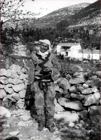 Persona con traje tradicional de pie junto a un muro de piedras, con casas rurales y una montaña de fondo en blanco y negro