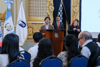 Hombre en podio, otro hombre de traje, mujer intérprete de señas, banderas de Guatemala, público de espalda, pared con molduras