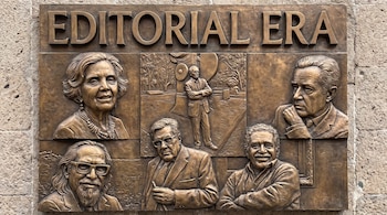 Placa de bronce en relieve con el texto EDITORIAL ERA, cuatro retratos de busto y una figura de cuerpo entero sobre fondo de paisaje en relieve, sobre una pared.
