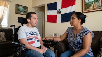 Un hombre joven en silla de ruedas motorizada habla con una mujer sentada a su lado en una sala. Al fondo, una bandera de la República Dominicana y cuadros.