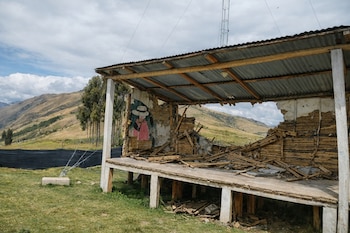 Imagen de un escenario o estructura exterior parcialmente colapsada, con escombros de madera y muros derrumbados. Hay un mural en la pared restante. Fondo de montañas y cielo nublado