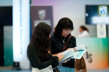 Dos mujeres leen un folleto abierto en un espacio con paneles informativos de fondo