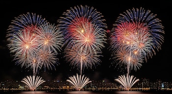 Tres grandes explosiones de fuegos artificiales rojos, azules y blancos sobre un cuerpo de agua con una ciudad iluminada en el horizonte.