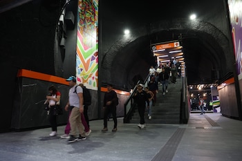 Vista interior de una estación de metro renovada con escaleras ascendentes y descendentes, pasajeros caminando, iluminación moderna y un mural colorido en la pared