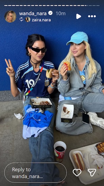 Dos mujeres con camisetas de Argentina y jeans sentadas en el suelo, comiendo sándwiches. Hay cajas de comida, una bandera, teléfono, auriculares y hot dogs