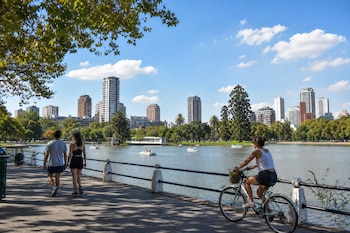 Personas caminan y andan en bicicleta por un paseo arbolado junto a un lago con botes y edificios en el fondo.