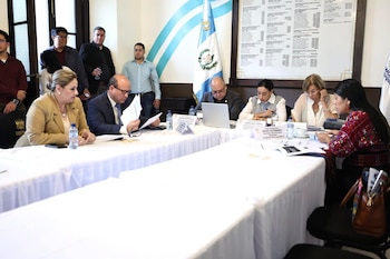 Personas reunidas alrededor de una mesa con documentos y computadoras portátiles frente a la bandera de Guatemala
