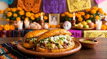 Dos pambazos con chorizo, papa y queso en un plato de barro, con un altar de flores de cempasúchil, velas y papel picado al fondo.