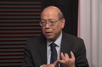 Un hombre asiático calvo con gafas, traje oscuro y camisa a cuadros, gesticula con las manos frente a un fondo de paneles acústicos negros y una pared clara