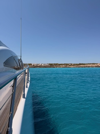 Lateral de un yate blanco con barandales, antena y cubierta de madera, navegando en un mar turquesa. Al fondo, costa árida con casas blancas