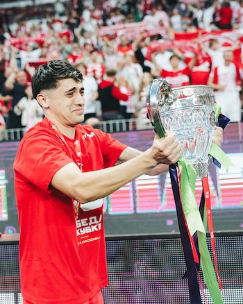 Un hombre con camiseta roja sostiene un trofeo de cristal y plata con cintas de colores. El trofeo tiene una parte separada, sugiriendo que está roto. Al fondo, una multitud borrosa