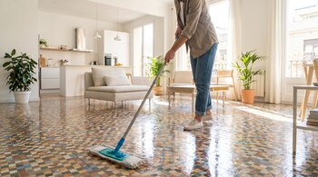 Una persona, vestida con jeans y camisa clara, trapea un suelo de mosaico brillante en una estancia moderna y luminosa con grandes ventanales y plantas.