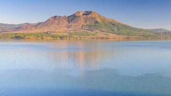 Um lago azul claro e escuro com pequenos barcos e remadores vistos de cima do mar, e um grande vulcão marrom e verde ao fundo sob um céu claro no Lago Malawi