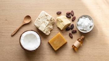 Vista superior de manteca de karité, cera de abeja, medio coco, granos de cacao, aceite de coco, frasco de aceite y una cuchara de madera.