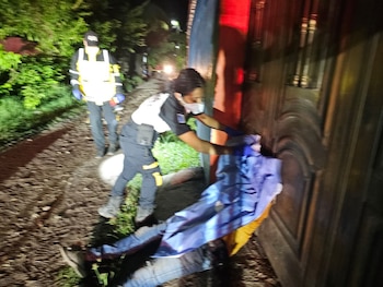 Dos personas con uniforme de emergencia y una con mascarilla atienden a un individuo tendido en el suelo en la noche, cerca de una puerta de madera y vegetación