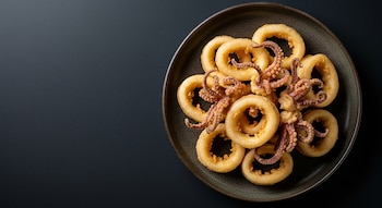 Imagen cenital de un plato oscuro con Kalamarakia tiganita: aros de calamar fritos dorados y tentáculos sobre un fondo oscuro y liso.