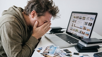 Hombre con cabello revuelto y sudadera gris cubre su rostro con las manos frente a una laptop con pantalla "Storage Full", rodeado de discos duros y tarjetas SD.