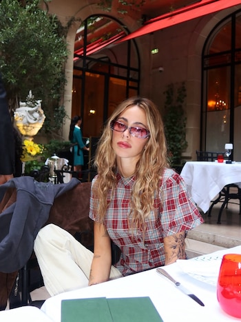 Una mujer con cabello largo rubio, lentes de sol y camisa a cuadros se sienta en una mesa de restaurante, con plantas y edificios de fondo