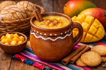 Un tazón de atole de mango humeante con trozos de fruta y canela, dos mangos enteros, un mango cortado, mango en cubos, galletas y conchas.