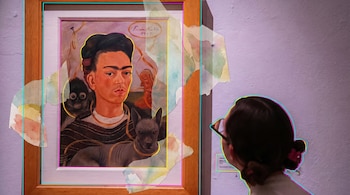 Una pintura enmarcada de Frida Kahlo, un autorretrato con un mono y un perro, observada por una persona de espaldas en una galería de arte con paredes moradas.