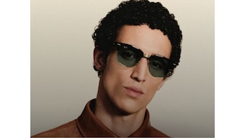 Retrato de un hombre con cabello rizado que lleva lentes de sol con marco negro y dorado sobre un fondo gris claro
