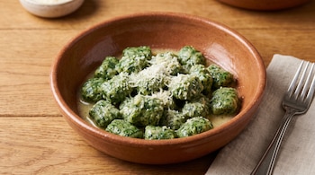 Cuenco de barro con ñoquis verdes y queso rallado, acompañado de un tenedor sobre una servilleta en una mesa de madera.