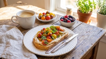 Una tostada con tomates cherry sobre un plato blanco, una taza de café, un tazón con frutos rojos y plantas junto a una ventana.