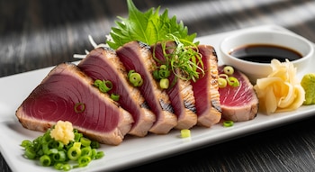 Primer plano de tataki de atún en un plato blanco, con seis rebanadas rosadas con bordes sellados, decoradas con cebolletas y jengibre, junto a salsa de soja.