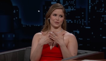 Erin Moriarty sentada en un sofá gris con un vestido rojo sin tirantes y un collar de diamantes, sus manos juntas sobre el pecho, cabello castaño