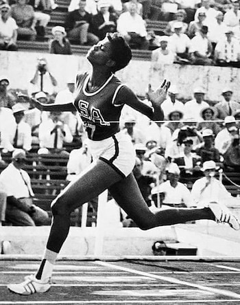 Fotografía en blanco y negro de una mujer atleta corriendo en una pista. Viste camiseta con las letras USA y shorts. El público se sienta en las gradas traseras