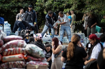 Un grupo de personas jóvenes maneja pilas de botellas de agua y bolsas de suministros al aire libre, con follaje verde de árboles de fondo