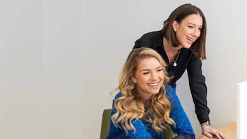 Dos mujeres sonrientes, una rubia de cabello largo y otra de pelo corto oscuro, miran hacia abajo en un gesto colaborativo frente a una pared clara