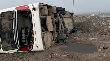 Un bus blanco de pasajeros está volcado sobre su lado izquierdo en un terreno árido junto a una carretera. El cielo es brumoso y gris