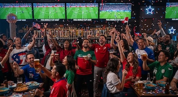 Multitud de personas en un bar de Dallas con los brazos en alto, gritando de alegría bajo una lluvia de confeti, mientras ven un partido de fútbol en grandes pantallas.