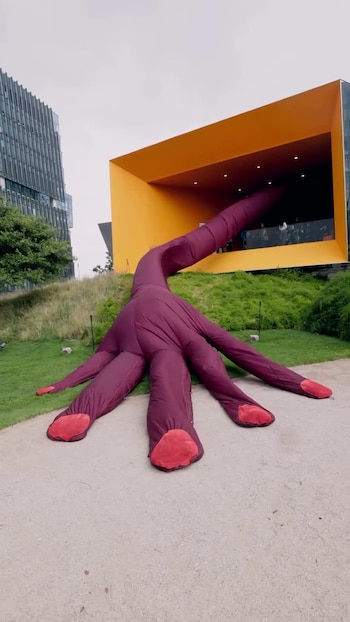 Escultura inflable de color púrpura y rojo con forma ramificada sobre césped y camino pavimentado, frente a un edificio con una apertura naranja y otro de cristal