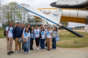 Once estudiantes diversos posan sonrientes frente a un transbordador espacial y un avión de prueba blanco de NASA en un museo al aire libre