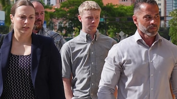 Timothy Hudson, un joven rubio con camisa gris, camina al aire libre entre dos adultos; una mujer con blazer azul y un hombre con camisa clara