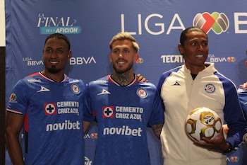 Tres hombres de pie: dos con camisetas azules de Cruz Azul y uno con una chaqueta blanca y azul, sosteniendo un balón de fútbol, ante un fondo azul con logos de Liga BBVA MX