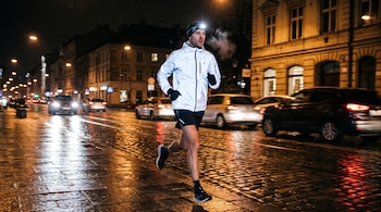 Hombre corriendo de noche en una calle empedrada mojada, lleva chaqueta reflectante, pantalones cortos, linterna frontal y guantes; se ve su aliento.