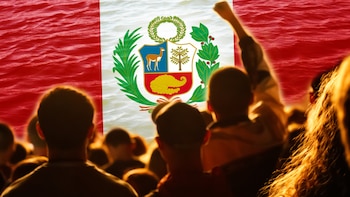 Multitud de personas vistas de espalda y en contraluz. Al fondo se observa una bandera de Perú, con bandas rojas laterales, banda blanca central y escudo nacional. Un puño en alto