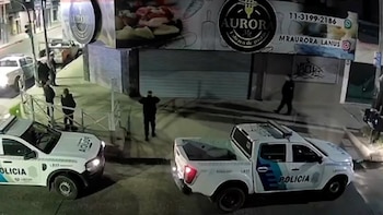 Vista aérea de dos camionetas policiales blancas y azules en una calle de noche, con varios oficiales y personas cerca de un edificio comercial cerrado