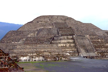 Vista frontal de la Pirámide del Sol en Teotihuacán, una gran estructura escalonada de piedra en tonos grises y ocres, bajo un cielo claro con montañas al fondo
