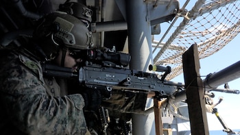 Un militar con casco y gafas de sol apunta una ametralladora montada. Al fondo se ven estructuras metálicas de un barco y una red protectora bajo un cielo azul