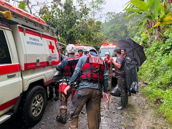 Tres rescatistas con uniformes de la Cruz Roja, cascos y chalecos, junto a un vehículo de rescate blanco. El fondo muestra un paisaje montañoso y húmedo con vegetación