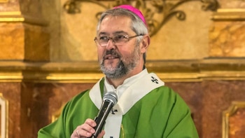 Un hombre con anteojos, solideo rosado y casulla verde sostiene un micrófono frente a un altar decorado