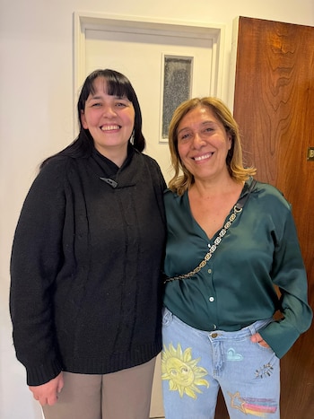 Dos mujeres sonríen de pie. Una tiene cabello oscuro, suéter negro y pantalones beige. La otra tiene cabello rubio, blusa verde y jeans con dibujos