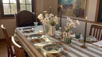 Mesa vestida con mantel a rayas, platos ilustrados, copas de vidrio verde, floreros con margaritas blancas y velas encendidas.